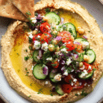 Loaded hummus platter topped with cucumber tomatoes feta olives and fresh herbs
